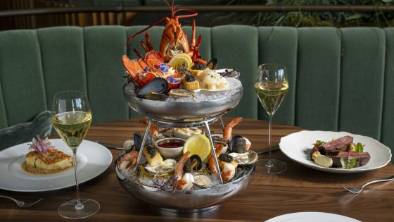An extravagant tiered seafood tower with a whole cooked lobster on top, surrounded by oysters, mussels, shrimp, scallops, and caviar, flanked by two glasses of champagne and two plated entrees on a wooden table.